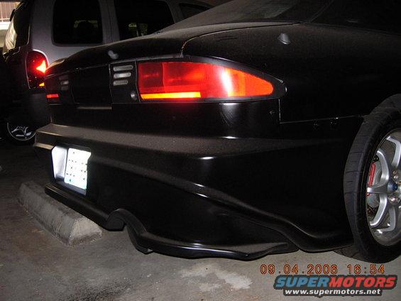 Rear view of a black car with a custom bumper and aftermarket taillight. Another vehicle is partially visible to the left