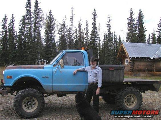 Man in cowboy hat and blue shirt leans on side of light blue pickup truck with flatbed and large tires. Black dog sits in front of truck. Log cabin and pine trees in background