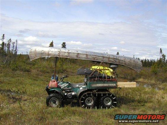 Green ATV with tracks, carrying a canoe and lumber in a grassy, wooded area