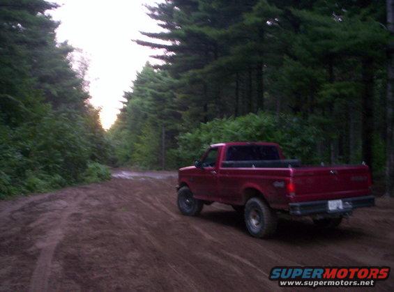 Red 1994 Ford F-150 pickup truck driving on a dirt road through a forest