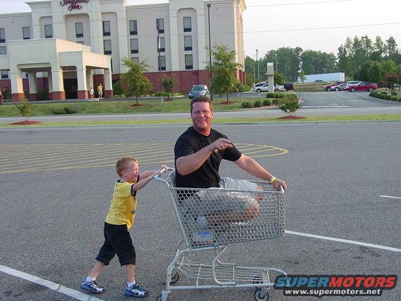 Man sits in shopping cart while a boy pushes him across a parking lot in front of a hotel