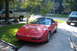 1994 Chevrolet Corvette - A red convertible sports car is parked in a driveway on a sunny day, with two trucks visible in the background.