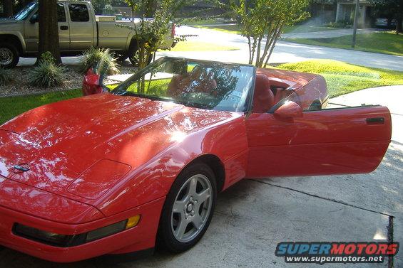 Red convertible sports car with doors open, parked on a driveway. A pickup truck is visible in the background