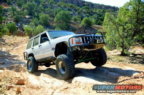 White Jeep Cherokee XJ with large tires and a brush guard driving on a dirt trail with trees and hills in the background