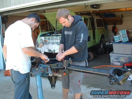 Two guys working on a green Ford Bronco in a garage. They are focused on a disassembled axle