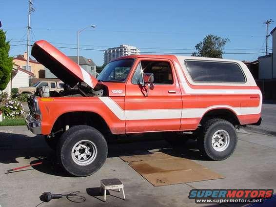 Orange and white Ford Bronco with hood up, parked on concrete. Tools and a step stool are nearby