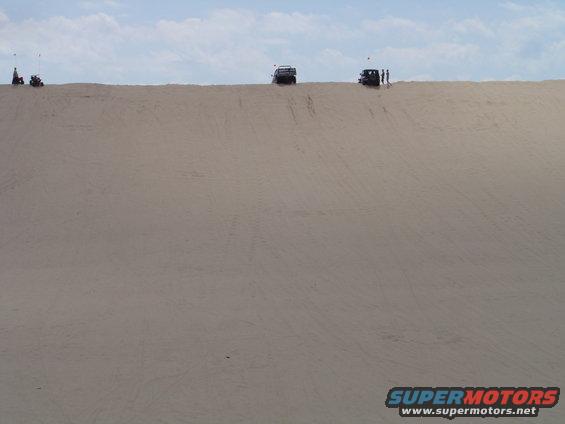 Several off-road vehicles and people on top of a large sand dune under a cloudy sky