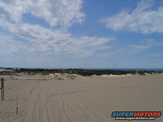 Sand dunes with a line of posts leading towards the ocean under a cloudy blue sky