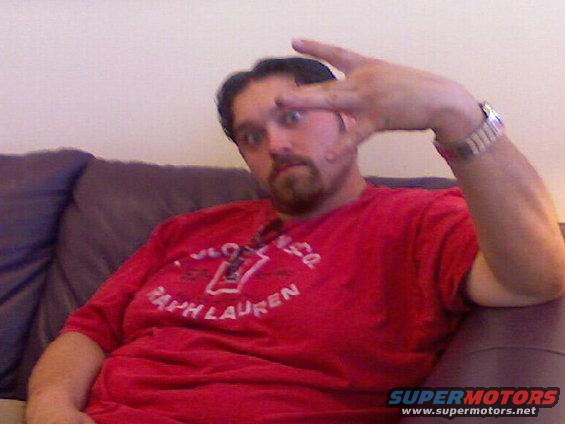Man with beard and goatee wearing a red Ralph Lauren t-shirt and a watch, making a peace sign