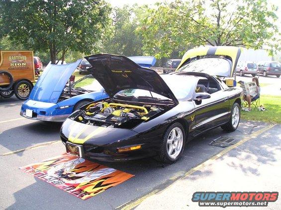 Black 1994 Pontiac Trans Am with hood open, yellow engine accents. Blue car with hood open behind it. Hartford Sign wooden display on left