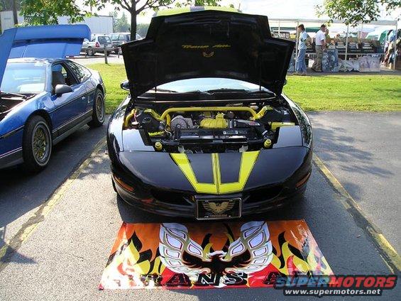 Black Pontiac Trans Am with yellow accents, hood open showing engine. Blue Pontiac Fiero parked to the left. Display mat in front of Trans Am