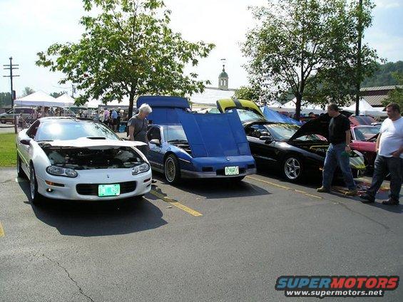 White Camaro, blue Fiero, black Trans Am, and pink car at outdoor event. Hoods are open on most