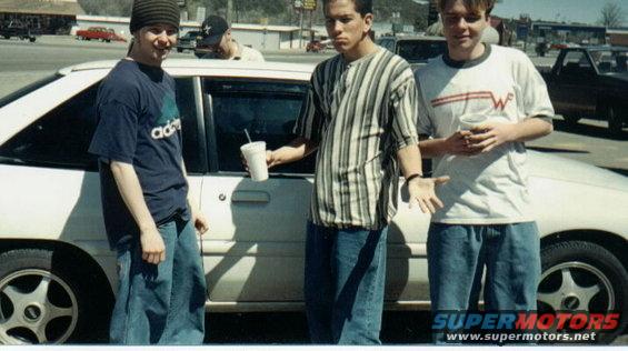 Three young men stand by a white car, one holding a drink. Another car is visible in the background
