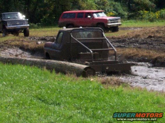 Modified pickup truck stuck in mud pit, another pickup and red SUV in background