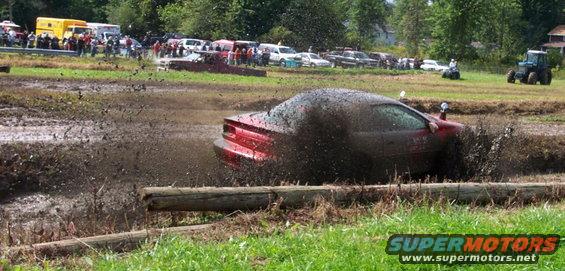 Red sports car stuck in a mud bog with spectators and other vehicles in the background
