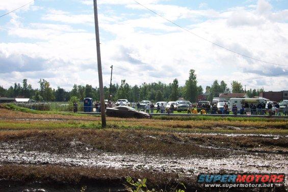 Muddy field with parked cars and spectators under a cloudy sky
