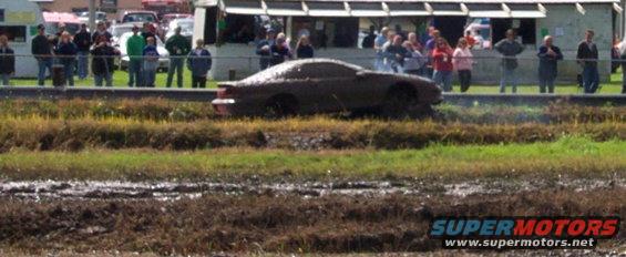 Mud-covered car stuck in a muddy field with spectators watching