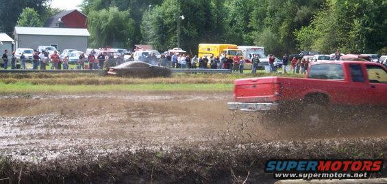 Red pickup truck kicking up mud, brown car in background, spectators watch mud race