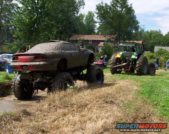Mud-covered car on monster truck wheels, towed by tractor