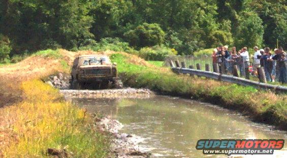Muddy 1978 Ford Bronco driving through a water obstacle with spectators on the bank