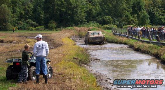 Truck driving through a mud bog with spectators watching from a fence line