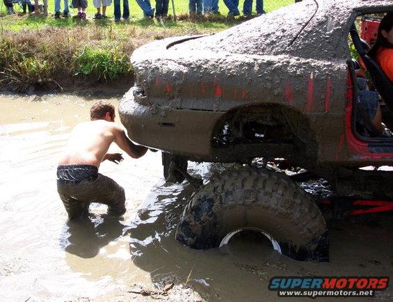 Mud-covered red car with large tires stuck in water, a person is checking underneath