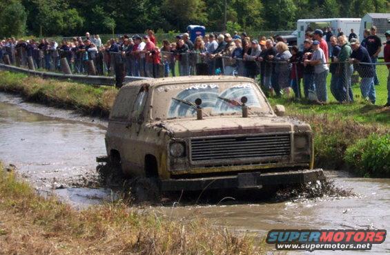 Muddy SUV driving through a mud pit with spectators watching