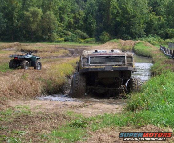 Muddy truck driving through water, ATV on grassy bank nearby