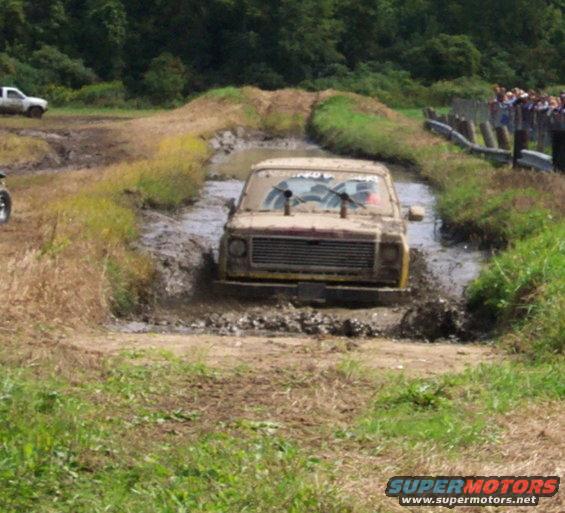 Mud-splattered 1970s pickup truck with dual exhaust stacks driving through a mud pit