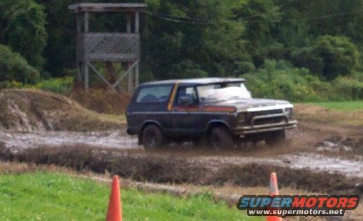 Dark blue Ford Bronco driving through mud, with a wooden tower in the background