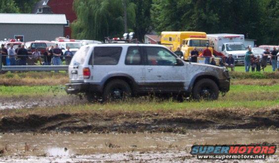 White SUV stuck in mud, with emergency vehicles and spectators in the background