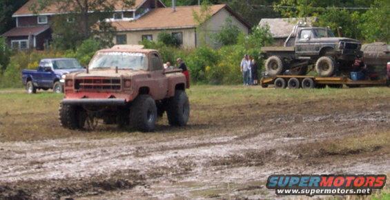 Mud-covered red pickup truck with oversized tires and a lifted suspension, viewed from the front