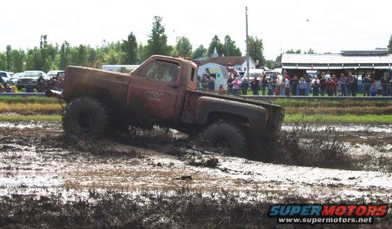 Red pickup truck with large tires stuck in mud pit, spectators in background