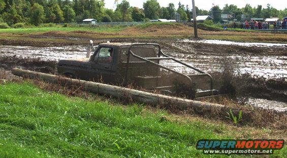 Muddy pickup truck with roll cage stuck in a mud pit, log in foreground