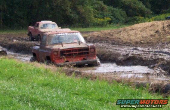 Two red trucks stuck in a mud pit with grassy banks
