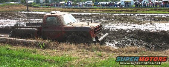 Red pickup truck stuck in mud, front right wheel submerged, exhaust pipes visible