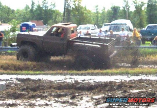 Muddy pickup truck with large tires in a mud bog, other vehicles and spectators in background