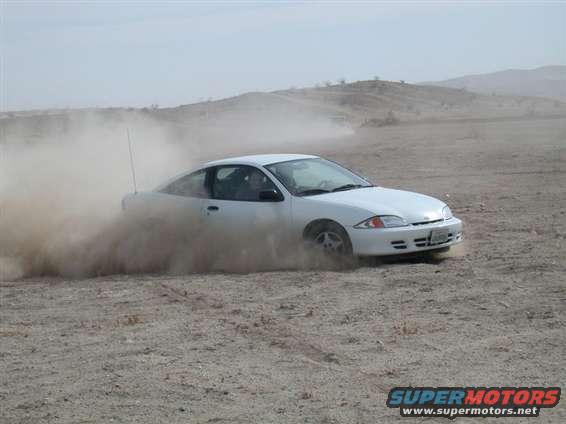 White coupe drifting in dirt, kicking up a large dust cloud. Appears to be a 1995 Chevrolet Cavalier