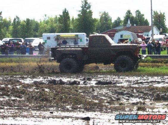 Mud-covered pickup truck with large tires driving through mud, side view