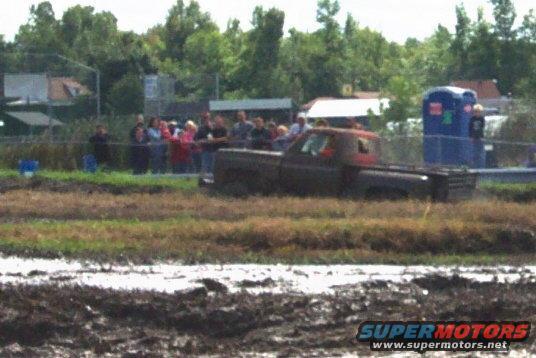 Pickup truck stuck in mud pit with spectators watching. Portable toilet in background