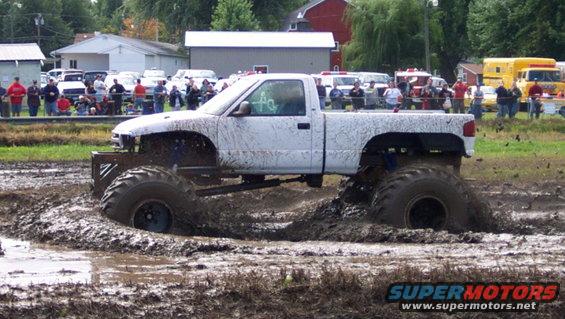 White pickup truck with large tires stuck in a mud pit, spectators in background