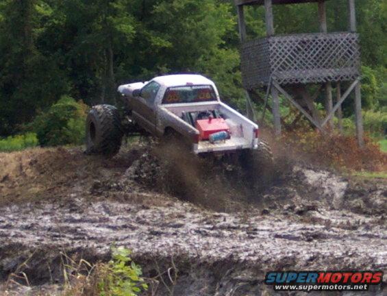 Monster truck with large tires driving through mud, kicking up dirt. A red gas can is in the truck bed