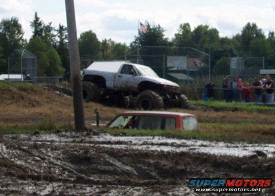 White lifted pickup truck with large tires on muddy hill, red SUV submerged in mud pit below