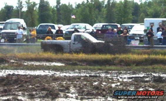 White pickup truck stuck in mud pit with spectators in background