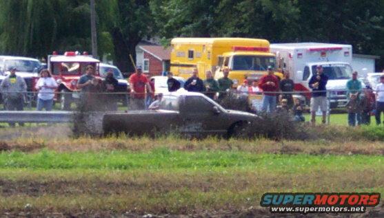 Pickup truck stuck in mud with spectators watching; emergency vehicles in background