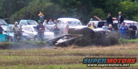 Mud bogging vehicle with engine exposed, kicking up mud, with spectators and other vehicles in background