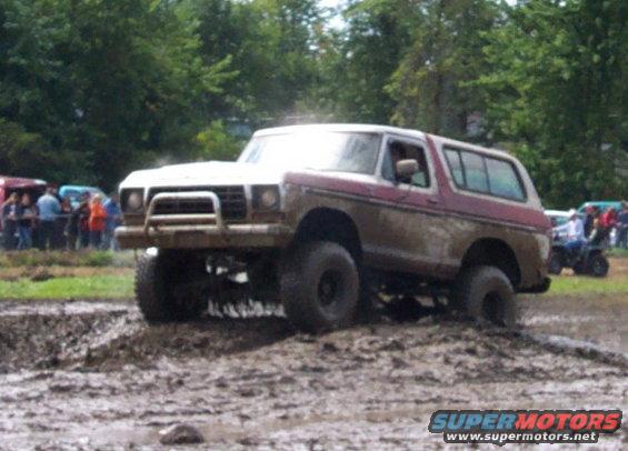 Muddy SUV with lifted suspension driving through a mud pit. Other vehicles and people in background