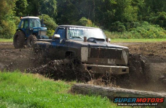 Mud-splattered black pickup truck with a hood scoop driving through mud, blue tractor in background