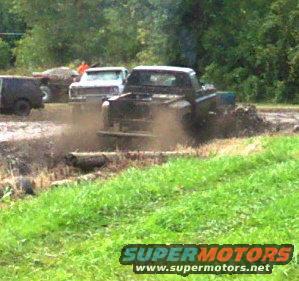 Several trucks driving through a mud bog with grass and trees in the background