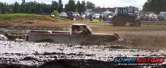 Pickup truck driving through a mud bog with a tractor and spectators in the background